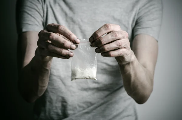 A luta contra as drogas e o tema da toxicodependência: viciado segurando pacote de cocaína em uma camiseta cinza em um fundo escuro no estúdio — Fotografia de Stock