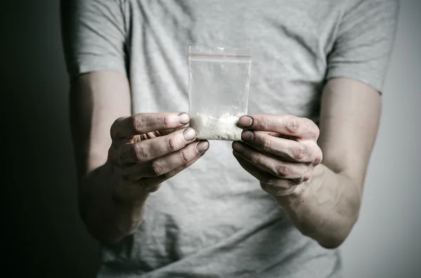 A luta contra as drogas e o tema da toxicodependência: viciado segurando pacote de cocaína em uma camiseta cinza em um fundo escuro no estúdio — Fotografia de Stock