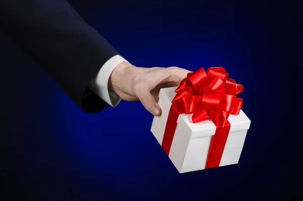 Il tema delle celebrazioni e dei regali: un uomo in abito nero con in mano un regalo esclusivo avvolto in una scatola bianca con nastro rosso, bellissimo e costoso regalo su uno sfondo blu scuro in studio isolato — Foto Stock