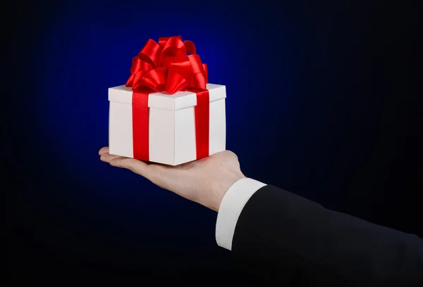 Le thème des célébrations et des cadeaux : un homme en costume noir tenant un cadeau exclusif enveloppé dans une boîte blanche avec ruban rouge, beau et cher cadeau sur un fond bleu foncé en studio isolé — Photo