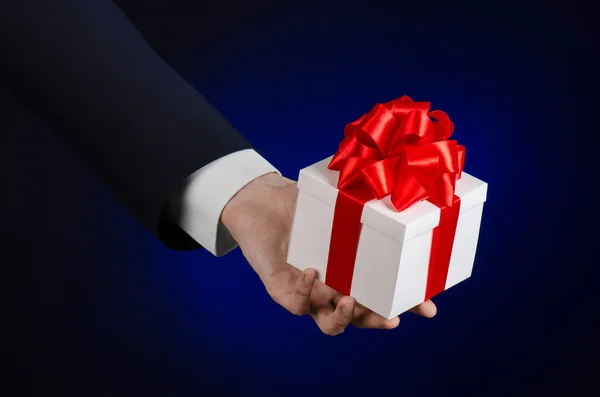 Il tema delle celebrazioni e dei regali: un uomo in abito nero con in mano un regalo esclusivo avvolto in una scatola bianca con nastro rosso, bellissimo e costoso regalo su uno sfondo blu scuro in studio isolato — Foto Stock