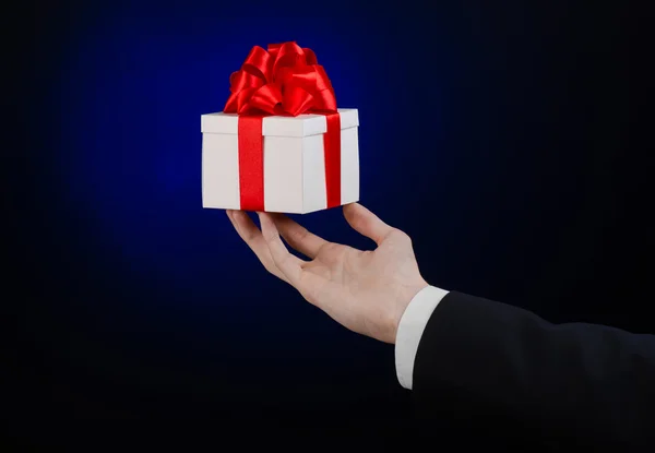 The theme of celebrations and gifts: a man in a black suit holding a exclusive gift wrapped in white box with red ribbon, beautiful and expensive gift on a dark blue background in studio isolated — Stock Photo, Image