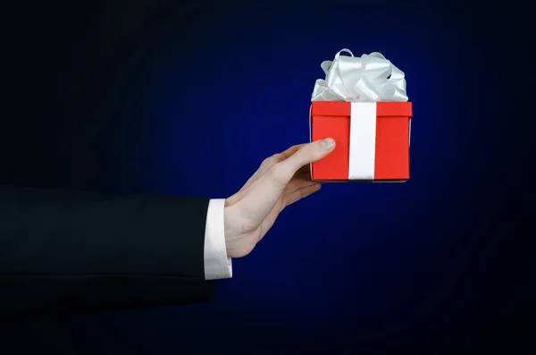 El tema de las celebraciones y regalos: un hombre en un traje negro con un regalo exclusivo envuelto en caja roja con cinta blanca, hermoso y caro regalo sobre un fondo azul oscuro en el estudio aislado — Foto de Stock