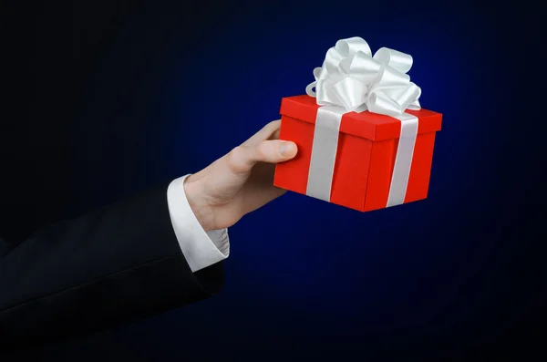 The theme of celebrations and gifts: a man in a black suit holding a exclusive gift wrapped in red box with white ribbon, beautiful and expensive gift on a dark blue background in studio isolated — Stock Photo, Image