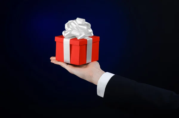 Le thème des célébrations et des cadeaux : un homme en costume noir tenant un cadeau exclusif enveloppé dans une boîte rouge avec ruban blanc, beau et cher cadeau sur un fond bleu foncé en studio isolé — Photo