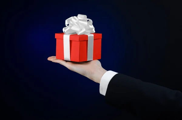 The theme of celebrations and gifts: a man in a black suit holding a exclusive gift wrapped in red box with white ribbon, beautiful and expensive gift on a dark blue background in studio isolated — Stock Photo, Image