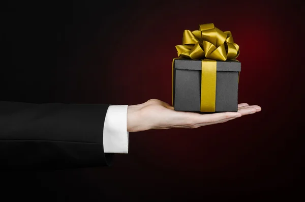 The theme of celebrations and gifts: a man in a black suit holding a exclusive gift packaged in a black box with gold ribbon, beautiful and expensive gift on a dark red background in studio isolated — Stock Photo, Image