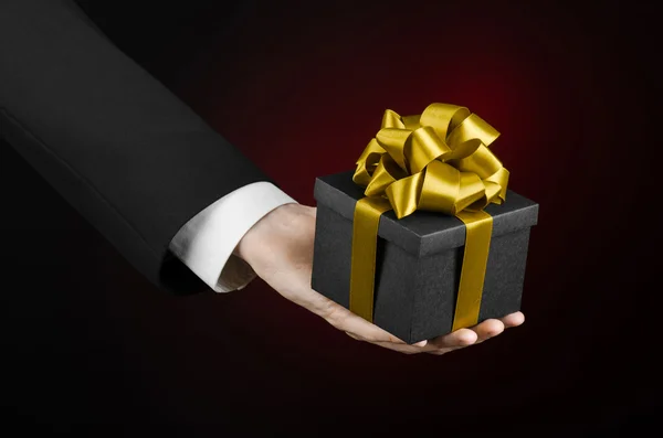 The theme of celebrations and gifts: a man in a black suit holding a exclusive gift packaged in a black box with gold ribbon, beautiful and expensive gift on a dark red background in studio isolated — Stock Photo, Image