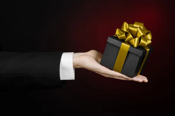 庆祝活动和礼品的主题: 一个穿着一套黑色的西装，深红色背景中分离的工作室举行独家礼品包装在一个黑箱与美丽的金色彩带和昂贵的礼物 — 图库照片