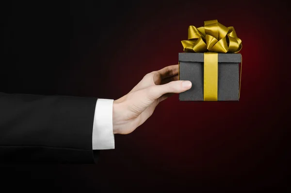 The theme of celebrations and gifts: a man in a black suit holding a exclusive gift packaged in a black box with gold ribbon, beautiful and expensive gift on a dark red background in studio isolated — Stock Photo, Image