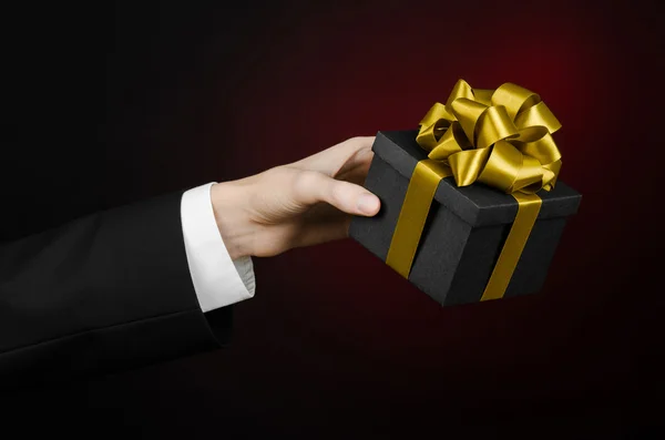 Le thème des célébrations et des cadeaux : un homme en costume noir tenant un cadeau exclusif emballé dans une boîte noire avec ruban d'or, beau et cher cadeau sur un fond rouge foncé en studio isolé — Photo