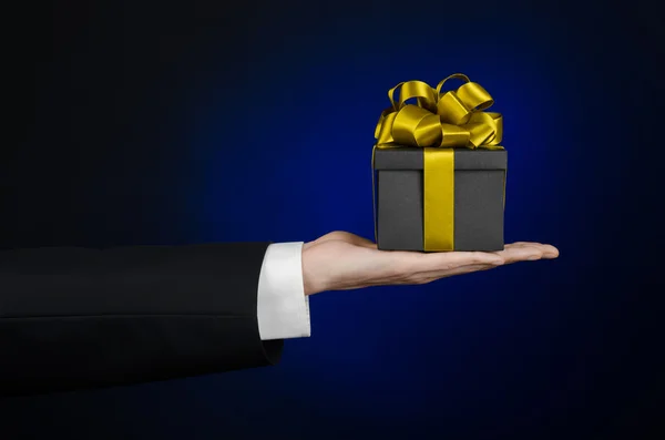 The theme of celebrations and gifts: a man in a black suit holding a exclusive gift packaged in a black box with gold ribbon, beautiful and expensive gift on a dark blue background in studio isolated — Stock Photo, Image