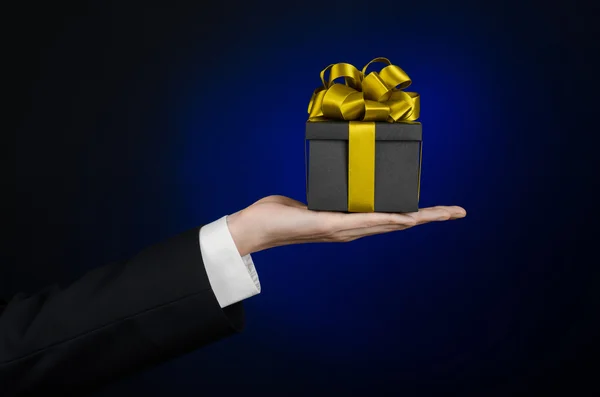 Das Thema der Feiern und Geschenke: ein Mann im schwarzen Anzug hält ein exklusives Geschenk in einer schwarzen Schachtel mit goldenem Band verpackt, schönes und teures Geschenk auf dunkelblauem Hintergrund im Studio isoliert — Stockfoto