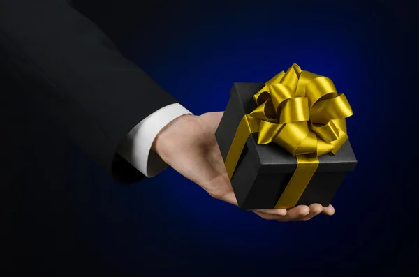 Das Thema der Feiern und Geschenke: ein Mann im schwarzen Anzug hält ein exklusives Geschenk in einer schwarzen Schachtel mit goldenem Band verpackt, schönes und teures Geschenk auf dunkelblauem Hintergrund im Studio isoliert — Stockfoto