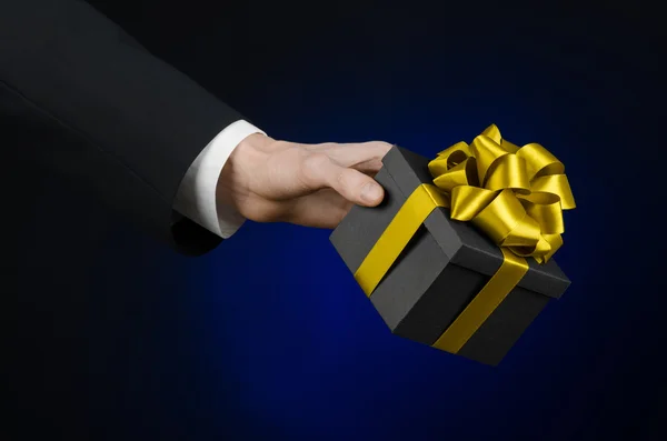 The theme of celebrations and gifts: a man in a black suit holding a exclusive gift packaged in a black box with gold ribbon, beautiful and expensive gift on a dark blue background in studio isolated — Stock Photo, Image