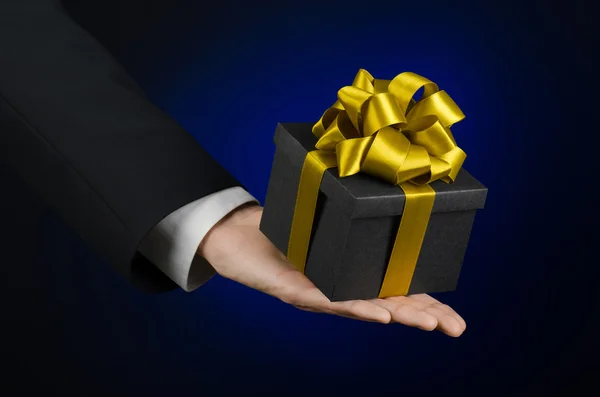 Le thème des célébrations et des cadeaux : un homme en costume noir tenant un cadeau exclusif emballé dans une boîte noire avec ruban d'or, beau et cher cadeau sur un fond bleu foncé en studio isolé — Photo