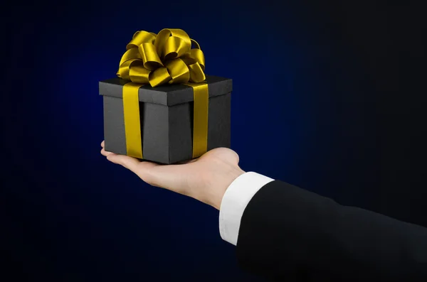 Kutlamalar ve hediye Tema: holding özel hediye altın şerit, güzel bir kara kutu ambalajlı ve pahalı hediye izole Studio koyu mavi zemin üzerine siyah takım elbiseli bir adam — Stok fotoğraf