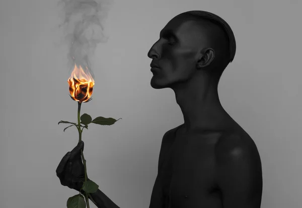 Gothic and Halloween theme: a man with black skin holding a burning rose, black death isolated on a gray background in studio — Stock Photo, Image