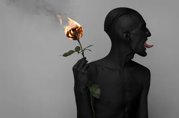 Tema gótico y de Halloween: un hombre con piel negra sosteniendo una rosa ardiente, la muerte negra aislada sobre un fondo gris en el estudio — Foto de Stock