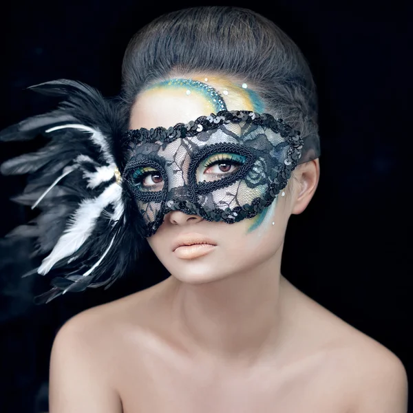 Geheimnisvolle schöne Mädchen in einer schwarzen Maske mit Federn mit grünen kreativen Make-up im Studio auf einem dunklen Hintergrund isoliert — Stockfoto