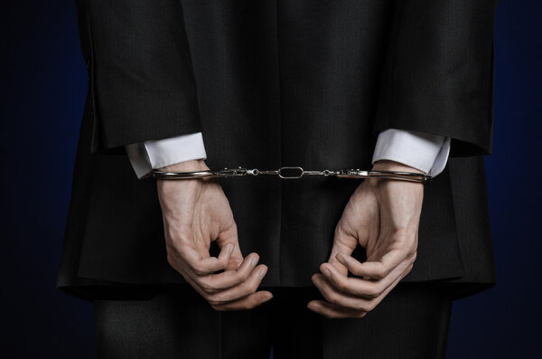 Коррупция и взяточничество тема: бизнесмен в черном костюме с наручниками на руках на темно-синем фоне в студии изолированный вид сзади
