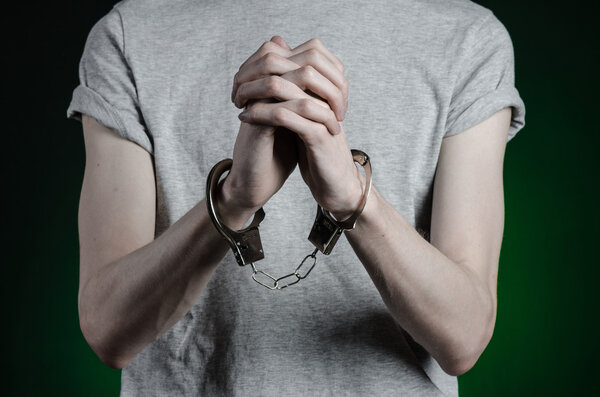 Prison and convicted topic: man with handcuffs on his hands in a gray T-shirt and blue jeans on a dark green background in the studio, put handcuffs on the drug dealer