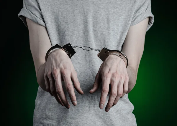 Prison and convicted topic: man with handcuffs on his hands in a gray T-shirt and blue jeans on a dark green background in the studio, put handcuffs on the drug dealer