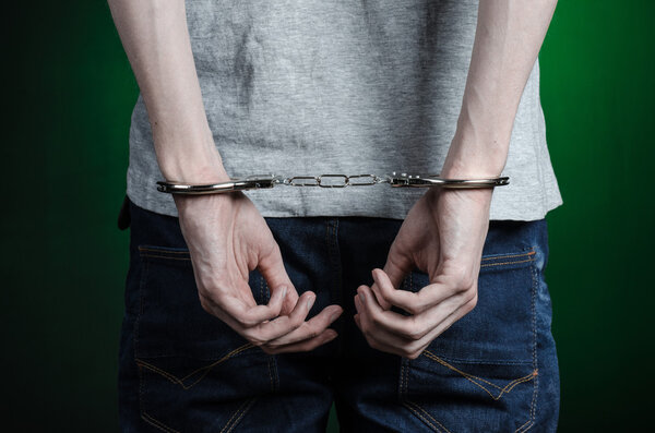 Prison and convicted topic: man with handcuffs on his hands in a gray T-shirt and blue jeans on a dark green background in the studio, put handcuffs on the drug dealer, the view from the back