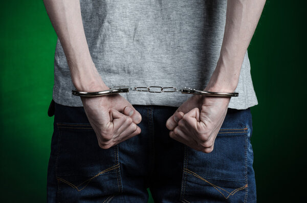 Prison and convicted topic: man with handcuffs on his hands in a gray T-shirt and blue jeans on a dark green background in the studio, put handcuffs on the drug dealer, the view from the back