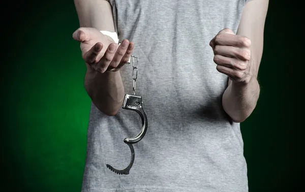 Gevangenis en veroordeelde onderwerp: man met handboeien op zijn handen in een grijs T-shirt en spijkerbroek op een donkere groene achtergrond in de studio, handboeien zetten de drugsdealer — Stockfoto