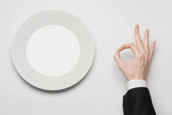 Business lunch and healthy food theme: man's hand in a black suit holding a white empty plate and shows finger gesture on an isolated white background in studio top view — Stock Photo, Image