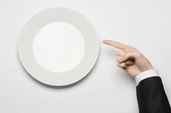 Business lunch and healthy food theme: man's hand in a black suit holding a white empty plate and shows finger gesture on an isolated white background in studio top view — Stock Photo, Image