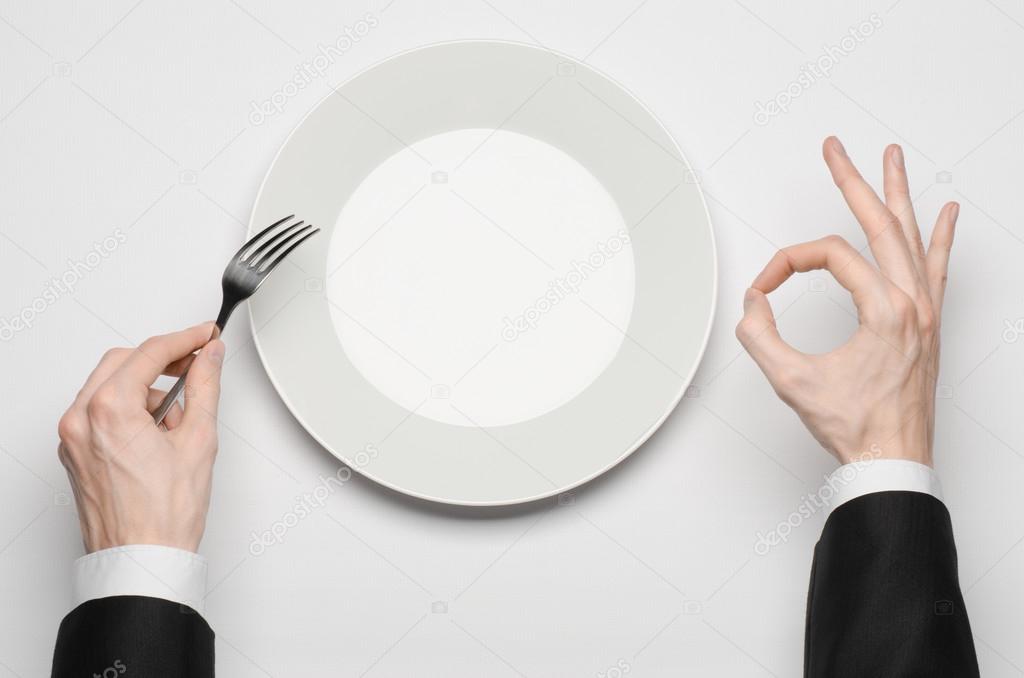 Business lunch and healthy food theme: man's hand in a black suit holding a white empty plate and shows finger gesture on an isolated white background in studio top view