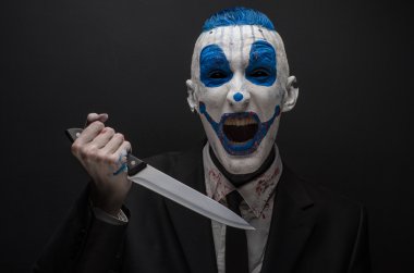 Terrible clown and Halloween theme: Crazy blue clown in a black suit with a knife in his hand isolated on a dark background in the studio