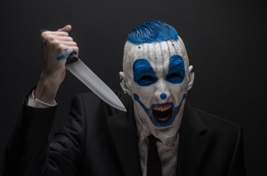 Terrible clown and Halloween theme: Crazy blue clown in a black suit with a knife in his hand isolated on a dark background in the studio