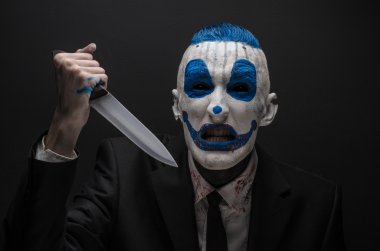 Terrible clown and Halloween theme: Crazy blue clown in a black suit with a knife in his hand isolated on a dark background in the studio