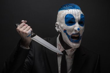 Terrible clown and Halloween theme: Crazy blue clown in a black suit with a knife in his hand isolated on a dark background in the studio