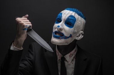 Terrible clown and Halloween theme: Crazy blue clown in a black suit with a knife in his hand isolated on a dark background in the studio