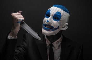 Terrible clown and Halloween theme: Crazy blue clown in a black suit with a knife in his hand isolated on a dark background in the studio