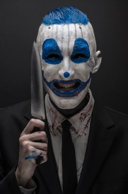Terrible clown and Halloween theme: Crazy blue clown in a black suit with a knife in his hand isolated on a dark background in the studio