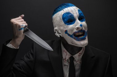 Terrible clown and Halloween theme: Crazy blue clown in a black suit with a knife in his hand isolated on a dark background in the studio