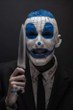 Terrible clown and Halloween theme: Crazy blue clown in a black suit with a knife in his hand isolated on a dark background in the studio