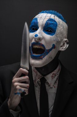 Terrible clown and Halloween theme: Crazy blue clown in a black suit with a knife in his hand isolated on a dark background in the studio