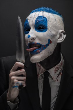 Terrible clown and Halloween theme: Crazy blue clown in a black suit with a knife in his hand isolated on a dark background in the studio
