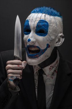 Terrible clown and Halloween theme: Crazy blue clown in a black suit with a knife in his hand isolated on a dark background in the studio