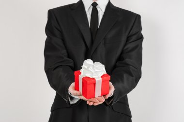 Theme holidays and gifts: a man in a black suit holds exclusive gift wrapped in red box with white ribbon and bow isolated on a white background in studio