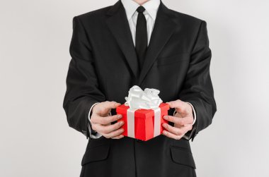 Theme holidays and gifts: a man in a black suit holds exclusive gift wrapped in red box with white ribbon and bow isolated on a white background in studio