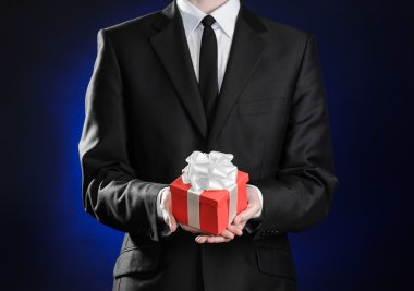 Theme holidays and gifts: a man in a black suit holds exclusive gift wrapped in red box with white ribbon and bow on a dark blue background in studio