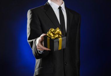 Theme holidays and gifts: a man in a black suit holds exclusive gift wrapped in a black box with gold ribbon and bow on a dark blue background in studio