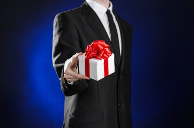 Theme holidays and gifts: a man in a black suit holds an exclusive gift in a white box wrapped with red ribbon and bow on a dark blue background in studio